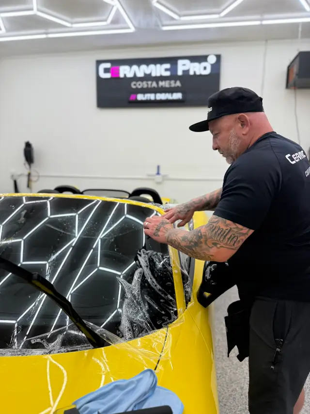 Installer in a Ceramic Pro shirt squeegeeing clear film onto the fender of a yellow car